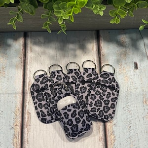 May include: Six grey and black leopard print keychains with silver key rings. Each keychain has a pocket for a small bottle of hand sanitizer.