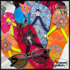 May include: A collection of colorful, decorative flip-flop eyeglass cases. The cases are in various colors, including red, orange, blue, and pink, and are embellished with floral and starfish designs. Some cases have the text "I CAN SEE CLEARLY NOW" embroidered on them. A pair of black eyeglasses rests in front.