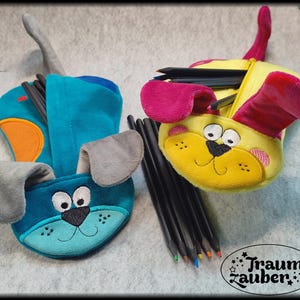 Könnte beinhalten: Zwei stiftförmige Federmäppchen in Hundeform, eines in Türkis und das andere in Gelb, jeweils mit Reißverschluss. Das türkisfarbene Etui hat graue Ohren und einen orangefarbenen Akzent, während das gelbe Etui rosa Ohren hat. Beide sind mit Stiften gefüllt. Der Text "Traumzauber" ist enthalten.