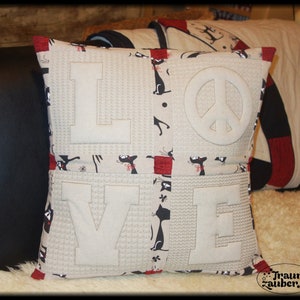 Op de afbeelding: Een beige kussen met het woord "LOVE" erop gestikt in wit. Het kussen is gemaakt van een patchwork stof met een zwart kattenontwerp en rode accenten. Een vredesteken is gestikt in het midden van het kussen.