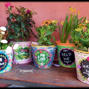 Könnte beinhalten: Fünf bunte Blumentöpfe mit gestickten und bemalten Botschaften. Die Töpfe sind mit Blumen verziert und tragen folgende Botschaften: "Ich werde überleben", "Halt an und riech an den Rosen", "Ich auch", "Hilf mir" und "Gieß mich".