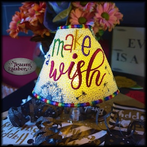 Könnte beinhalten: Ein weißer Lampenschirm mit einem Regenbogenrand und den Worten "make a wish" in bunten Fäden gestickt. Der Lampenschirm ist von innen beleuchtet und strahlt ein warmes Licht aus.