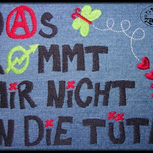 Puede incluir: Parche de mezclilla bordado con el texto "Das kommt mir nicht in die Tute" en letras negras. El texto está rodeado de bordados rojos, verdes y blancos. Hay una pequeña mariposa con alas verdes y cuerpo rojo en la esquina superior derecha. El texto "Traumzauber" está en la esquina superior derecha.
