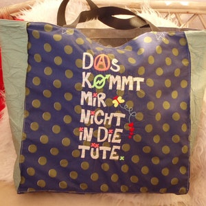 Op de afbeelding: Een blauwe en groene tote bag met witte stippen. De tas heeft een zwart handvat en de tekst "Das kommt mir nicht in die Tute" geborduurd op de voorkant.