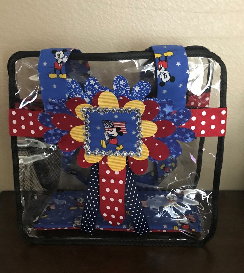 May include: Clear plastic tote bag with a red, white, and blue Mickey Mouse design. The bag has a black mesh pocket on the front and a red and white polka dot ribbon.