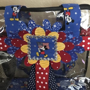 May include: A clear plastic tote bag with a red, white, and blue Mickey Mouse themed design. The bag has a large red, white, and blue flower with a Mickey Mouse image in the center. The bag also has red and white polka dot ribbon and blue ribbon with white stars.