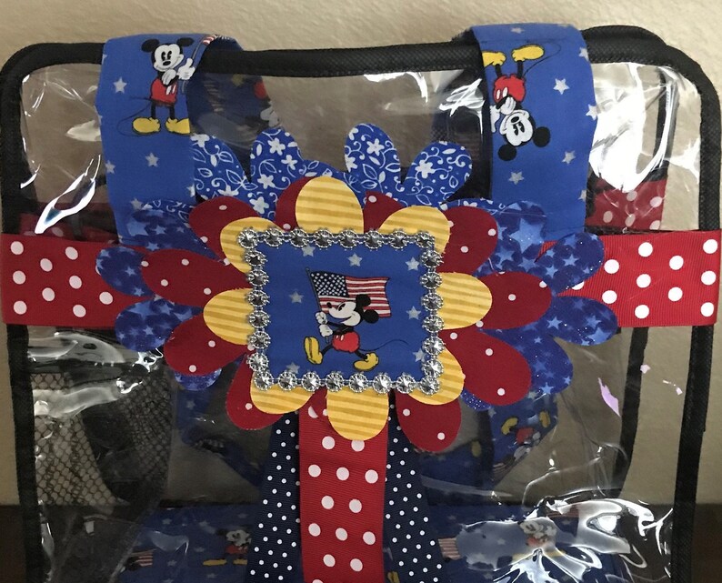 May include: A clear plastic tote bag with a red, white, and blue Mickey Mouse themed design. The bag has a large red, white, and blue flower with a Mickey Mouse image in the center. The bag also has red and white polka dot ribbon and blue ribbon with white stars.
