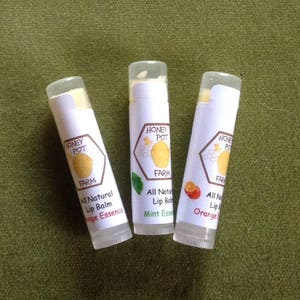 May include: Three lip balm tubes with white labels. The labels have a logo that says "Honey Pot Farm" with a lemon illustration. The lip balms are labelled "All Natural Lip Balm" with the flavours "Orange Essence", "Mint Essence", and "Orange".