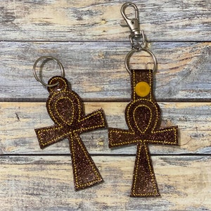 May include: Two brown glitter ankh keychains with yellow stitching. One keychain has a silver clasp, and the other has a silver keyring. The keychains are on a wooden background.