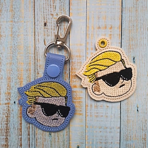 Könnte beinhalten: Zwei gestickte Schlüsselanhänger mit einer Comicfigur mit blonden Haaren und Sonnenbrille. Der Schlüsselanhänger links ist blau und der rechts ist beige.