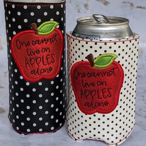 May include: Two black and white polka dot can coolers with red apple designs. The apple has a green leaf and the text "One cannot live on apples alone".