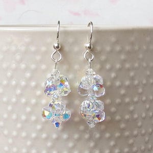 May include: A pair of clear crystal earrings with iridescent highlights. Each earring has a cluster of faceted beads hanging from a silver-coloured hook. The beads reflect a rainbow of colours, creating a sparkling effect. These earrings are a delicate and elegant accessory.