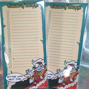 Peut inclure: Deux blocs-notes avec le texte "Cause of Death" et une illustration de dessin animé. Les blocs-notes ont des pages lignées et des cases à cocher. Un stylo blanc repose devant les blocs-notes. L'illustration comprend le texte "Better luck next time!"