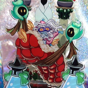 Peut inclure: Un présentoir en acrylique coloré représentant un personnage fantastique avec un manteau rouge, une barbe blanche et un visage bleu. Le présentoir comprend deux figurines en forme de potion avec du liquide bleu et des chapeaux noirs, et deux figurines vertes avec des yeux noirs. Le fond est irisé.