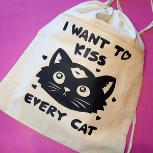 May include: A white canvas drawstring bag with black text that reads "I want to kiss every cat". A black cat with a heart-shaped nose is printed on the bag.