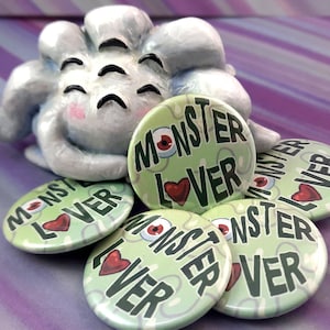 Puede incluir: Cinco botones de solapa verdes y blancos con el texto "Monster Lover" en negro y un corazón rojo. Los botones están dispuestos en círculo alrededor de un monstruo blanco, esponjoso y de tres ojos.