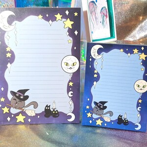 May include: Two blue and black Halloween themed note cards with a black cat wearing a witch's hat and a crescent moon with a face. The cards have a lined area for writing.