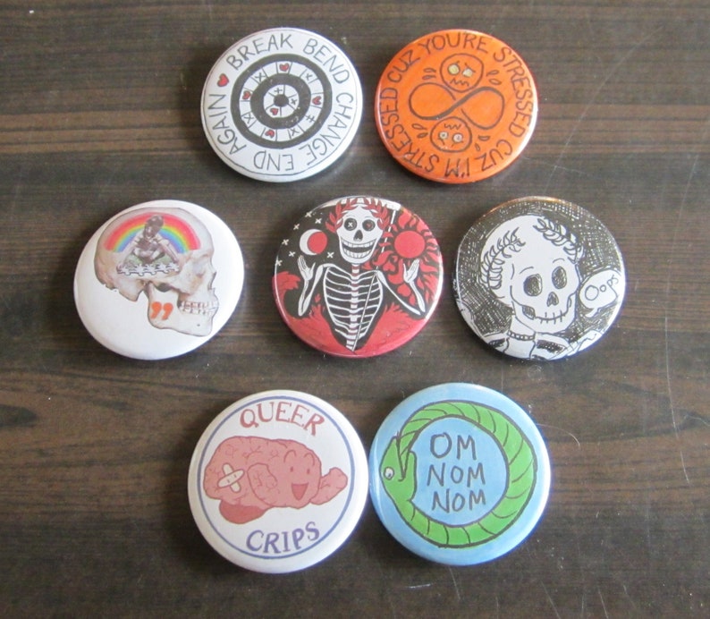 All seven button designs sitting in a flower shape. Break Bend Change End Again has the words around the edge, combined with looping ladders of hearts and Xs. Infinite Stress Loop is bright orange and shows a lemniscus and two stressed-out faces, surrounded by an infinite circle of text reading I'm Stressed Cuz You're Stressed Out Cuz... Skull collage is a photo of a skull, containing marker rainbows and a photo of a little girl making paper dolls. Susanna Jayne is in black, white, and red, showing a skeleton, wearing the laurels of triumph, holding the sun and the moon, erupting from an ocean of blood with a scythe in the background. Oops is black and white, showing a skeleton wearing the laurels of triumph and saying Oops. Queer Crips shows our little bandaged brain logo with the words Queer Crips around it. The Om Nom Nom Nouroboros shows a green ouroboros on a blue background, with the words OM NOM NOM at the center.