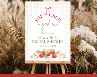 May include: A custom printed sign on an easel, featuring the text "She picked a good one" and "Welcome to Maggie's Bridal Shower, October 10, 2025." The sign has a gingham background and apple illustrations.