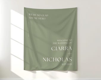 May include: Sage green wedding welcome banner with white text. The banner reads "WE'RE SO GLAD YOU'RE HERE!" and "WELCOME TO THE WEDDING OF CIARRA + NICHOLAS 11.23.2025". The banner is hanging on a white wall.