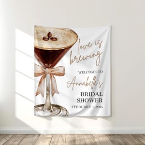 Op de afbeelding: Wit spandoek met een illustratie van een koffiecocktail en tekst. De tekst luidt "love is brewing", "Welcome to Annabelle's", "Bridal Shower" en "February 1, 2025". Het spandoek hangt aan een witte muur.