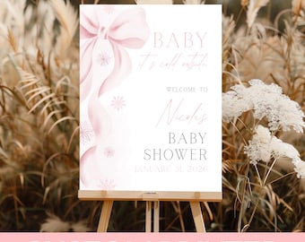 May include: A custom printed baby shower sign on an easel. The sign is white with pink accents, a bow, snowflakes, and the text "BABY It's cold outside. Welcome to Nicoli's BABY SHOWER January 31, 2026."