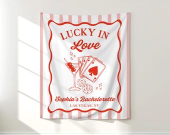 Pode incluir: Uma tapeçaria de parede listrada em branco e rosa com uma borda vermelha. A tapeçaria apresenta o texto "LUCKY IN Love" e um copo de martini, cartas de baralho e dados. O texto "Sophia's Bachelorette LAS VEGAS, NV" também está incluído.