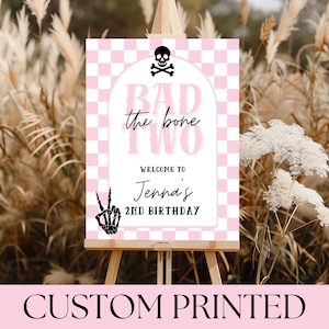 May include: A custom printed sign on an easel with a pink and white checkered background. The sign reads "BAD the bone TWO" with a skull and crossbones graphic. Below, it says "WELCOME TO Jenna's 2ND BIRTHDAY" with a skeleton hand making a peace sign.