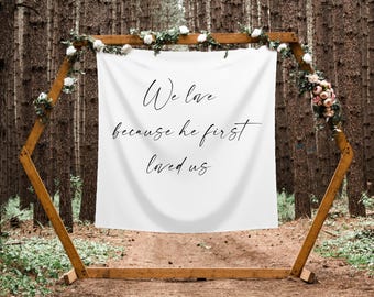 May include: White fabric banner with black calligraphy text that reads "We love because he first loved us" hanging from a wooden hexagonal archway decorated with white and pink flowers. The archway is in a forest setting.