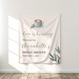 Op de afbeelding: Een crèmekleurige banner voor een bruidsdouche met de tekst "Love is brewing" en "Welcome to Annabelle's Bridal Shower February 1, 2025". De banner heeft een illustratie van een koffiekopje en groene bladaccenten.