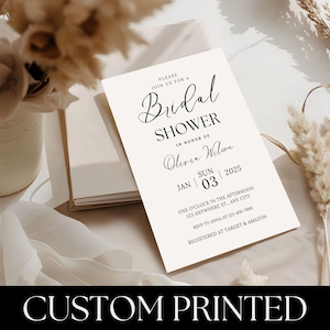 May include: A white card with black text announcing a bridal shower for Olivia Wilson on January 3, 2025, at one o'clock in the afternoon. The address is 123 Anywhere St., Any City. RSVP to Anna at 123-456-7890. The bride-to-be is registered at Target and Amazon.