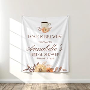 Op de afbeelding: Witte bruidsdouche banner met de tekst "LOVE IS BREWING" en "Welcome to Annabelle's Bridal Shower February 1, 2025". De banner heeft een koffiekopje afbeelding en bloemaccenten in bruin, oranje en geel.