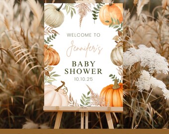 May include: A custom printed baby shower sign on an easel. The sign features watercolor pumpkins, greenery, and the text "Welcome to Jennifer's Baby Shower 10.10.25". The sign is set against a backdrop of tall, dried grasses.