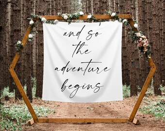 May include: White banner with black calligraphy that reads "and so the adventure begins" hanging from a wooden hexagonal archway decorated with greenery and flowers in a forest setting.