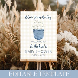 May include: A baby shower sign on an easel with the text "Blue Jean Baby." The sign features a denim pocket graphic with floral accents and the text "Welcome to Natalie's Baby Shower June 6, 2026." The background has a light beige gingham pattern.