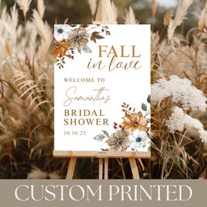 May include: A custom printed bridal shower sign on an easel. The sign features the text "Fall in love" and "Welcome to Samantha's Bridal Shower 10.10.25". Floral accents in brown, white, and orange decorate the sign.
