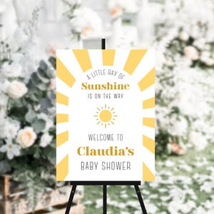 May include: A white sign with yellow sun rays and a yellow sun graphic. The sign says "A little ray of sunshine is on the way. Welcome to Claudia's Baby Shower."