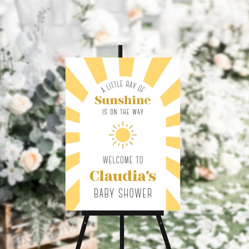 May include: A white sign with yellow sun rays and a yellow sun graphic. The sign says "A little ray of sunshine is on the way. Welcome to Claudia's Baby Shower."