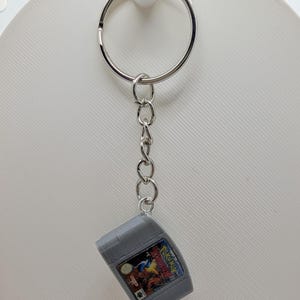 May include: A silver metal keychain featuring a miniature gray plastic replica of a Nintendo 64 game cartridge. The cartridge has a small rectangular label with the text Pokemon Stadium printed on it, attached to a circular metal ring by a chain.
