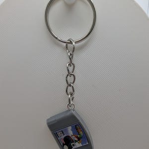 May include: A silver-colored metal keychain featuring a miniature gray plastic video game cartridge replica. The cartridge has a small rectangular label with a colorful graphic print and the text Nintendo 64 visible on the front surface.