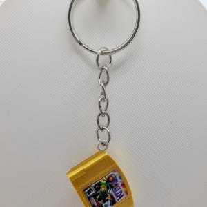 May include: A silver metal keychain featuring a miniature replica of a gold-colored video game cartridge. The cartridge displays a small label with the text 'ZELDA' and an 'E' rating symbol, attached to a standard circular split ring by a short chain.