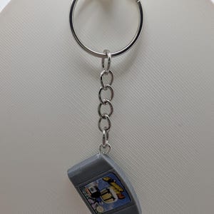 May include: A silver-toned metal keychain featuring a miniature replica of a grey video game cartridge. The cartridge has a rectangular label with the text "SUPER MARIO 64" printed in yellow and black, along with a small Nintendo logo.