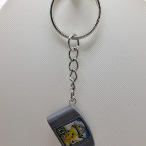 May include: A silver metal keychain featuring a miniature gray rectangular video game cartridge charm. The charm displays a small label with a yellow Pikachu character and the text Pikachu, along with an E rating symbol in the corner.