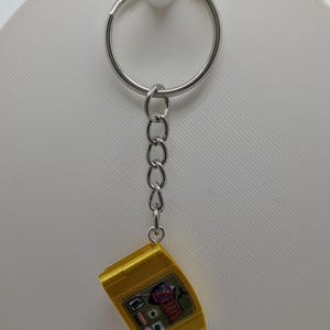 May include: A silver metal keychain featuring a miniature gold-colored video game cartridge replica. The cartridge has a label with the text The Legend of Zelda and an E rating symbol. The keychain includes a circular ring and a short chain link.