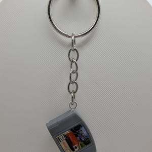 May include: A silver metal keychain featuring a miniature gray video game cartridge replica. The cartridge has a rectangular label with a colorful graphic of a red car and the text "Cruis'n USA" printed on the front, along with a small Nintendo logo.