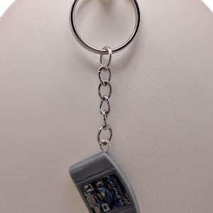 May include: A silver metal keychain featuring a miniature gray plastic video game cartridge replica. The cartridge has a rectangular label with blue and black graphics, a T rating symbol, and the text Star Fox 64 printed on the front surface.