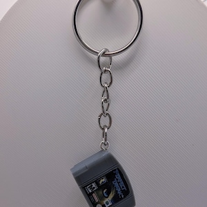 May include: A silver metal keychain featuring a miniature gray video game cartridge replica. The cartridge label displays the text Perfect Dark, an M rating symbol, and a Nintendo logo. The keychain includes a circular ring and a short chain link.