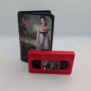 May include: A black wallet and a red VHS tape replica, both featuring the movie title "Jennifer's Body." The wallet displays a movie poster image. The VHS tape has a small image of the movie poster.