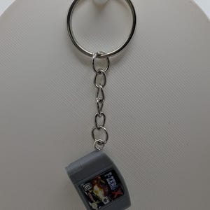 May include: A silver-toned metal keychain featuring a miniature gray plastic replica of an F-Zero X video game cartridge. The keychain includes a circular ring and a short chain attached to the top of the rectangular game cartridge, which displays the game title and an E rating symbol on its front label.