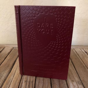 May include: A burgundy red hardcover book with a textured cover. The book has a circular design with the words "I DARE YOU!" in the center.